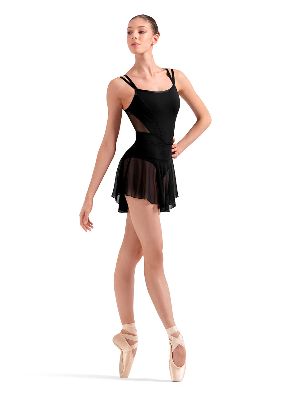 Dancer is wearing a mesh, pull-on ballet skirt with an elasticized waistband and high-low styling in Black. She has paired the skirt with a matching double spaghetti strap leotard, and pink pointe shoes, standing in a fourth position on pointe with her left arm on her hip.