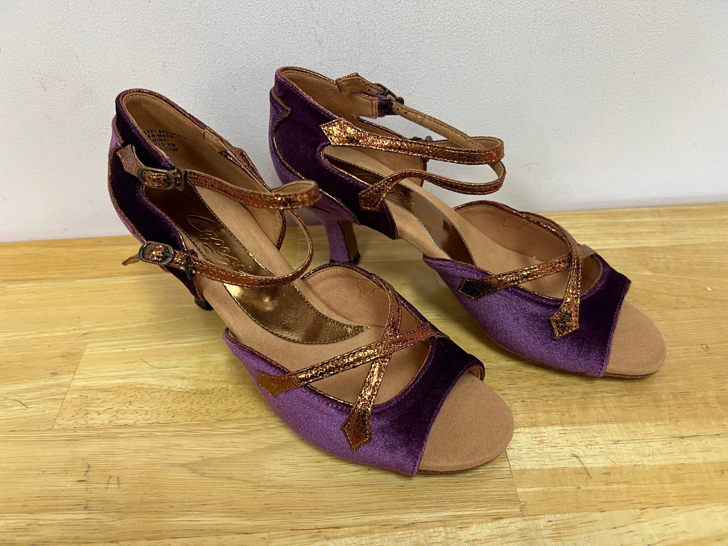 A  pair of Latin ballroom shoes with a 2" flared heel featuring a purple velvet vamp and heel with bronze trim. Side double buckle closure.