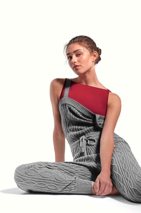 Dancer is wearing a black, tank strap, square neck, cable knit, warm -up jumpsuit with a tie front waist. Dancer has paired the garment with a red leotard and is sitting with her right leg tucked under her left.