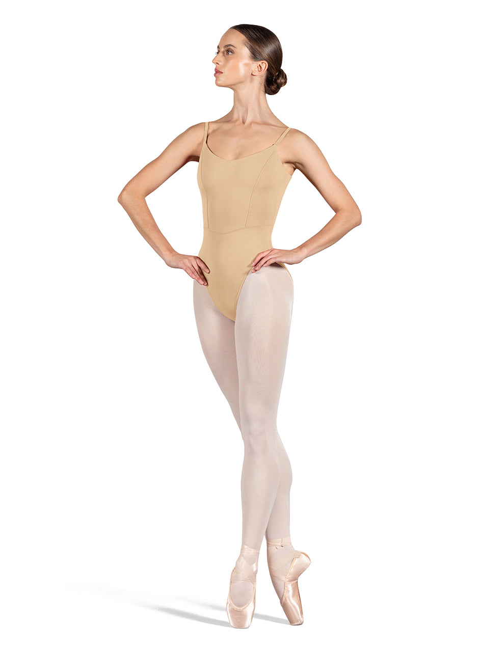 Dancer is wearing a sand, camisole leotard with a scoop neck and back and bodice seam detail. She has paired the garment with pink tights and pointe shoes and is balancing in a fourth position releve on pointe with her hands on her hips.