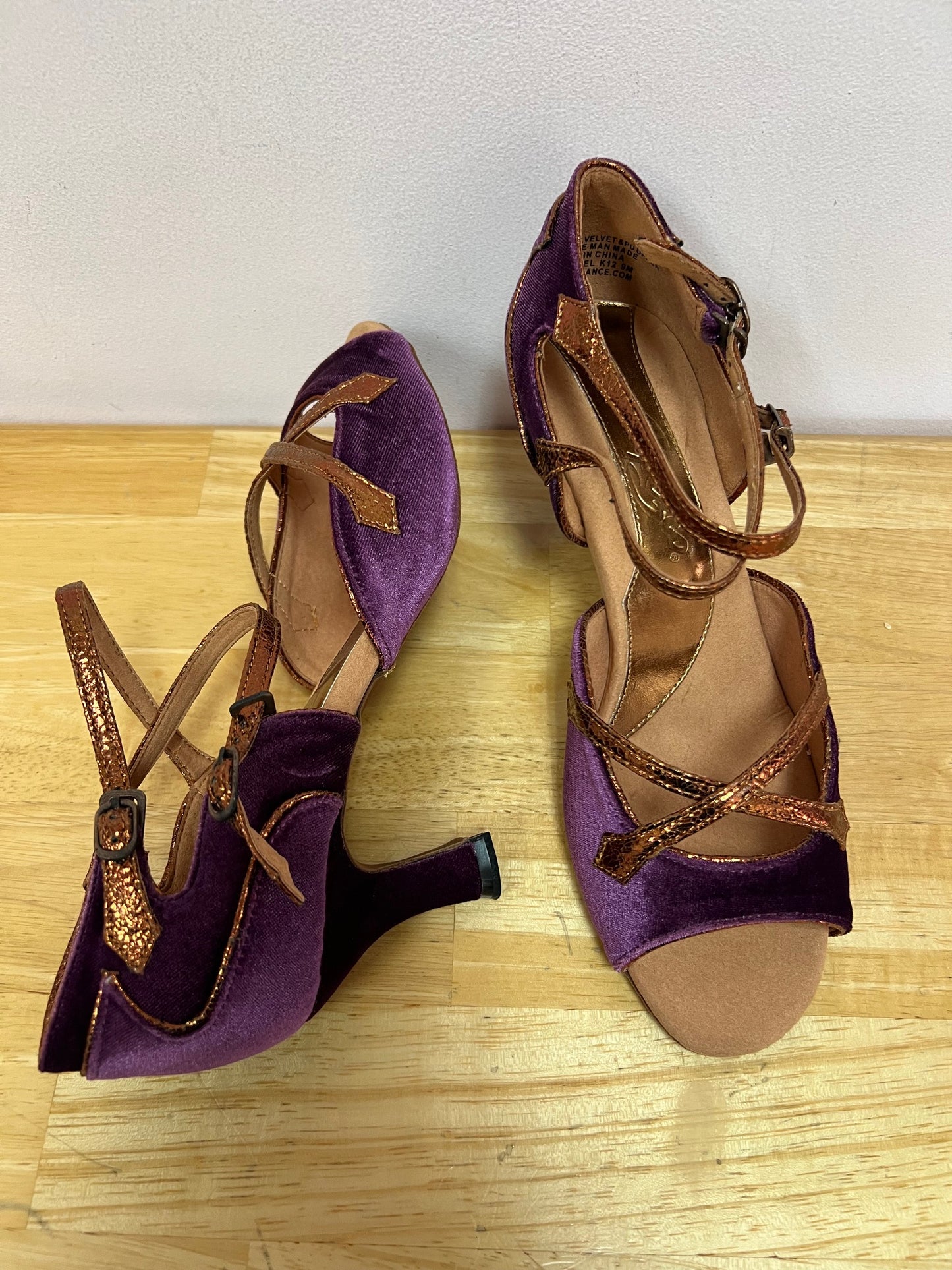 A  pair of Latin ballroom shoes with a 2" flared heel featuring a purple velvet vamp and heel with bronze trim. Side double buckle closure.