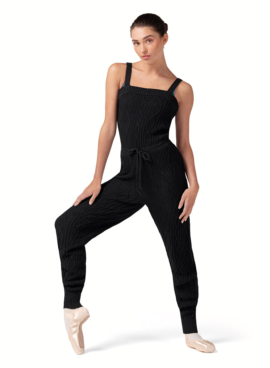 Dancer is wearing a black, tank strap, square neck, cable knit, warm -up jumpsuit with a tie front waist. Dancer has paired the garment with pink tights and pointe shoes and has her right foot on pointe and right hand on her leg.