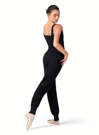 Dancer is wearing a black, tank strap, square neck, cable knit, warm -up jumpsuit with a tie front waist. Dancer has paired the garment with pink tights and pointe shoes and has her right foot pointed to the back and is looking over her right shoulder.