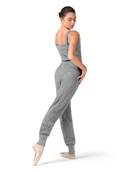 Dancer is wearing a grey marle, tank strap, square neck, cable knit, warm -up jumpsuit with a tie front waist. Dancer has paired the garment with pink tights and pointe shoes and has her right foot pointed to the back and is looking over her right shoulder.